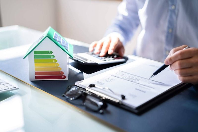 Imagen de una mesa de trabajo donde se ve una carpeta, una calculadora y una casa con el gráfico de eficiencia energética.